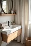 Scandinavian bathroom with a wood vanity, marble sink, black faucet, round mirror, and soft lighting from a window with curtains.