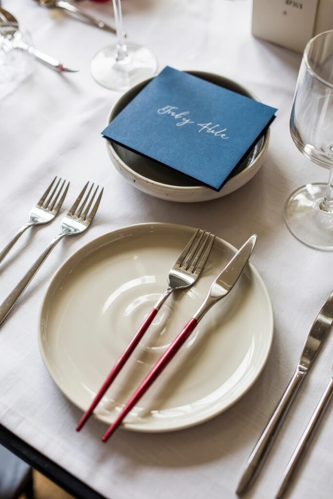 Patriotic 4th of July Table Décor & Centerpieces 2 Minimalist place setting with red-handled cutlery, white dinnerware, wine glasses, and a blue folded card reading “July 4th” placed in a bowl.