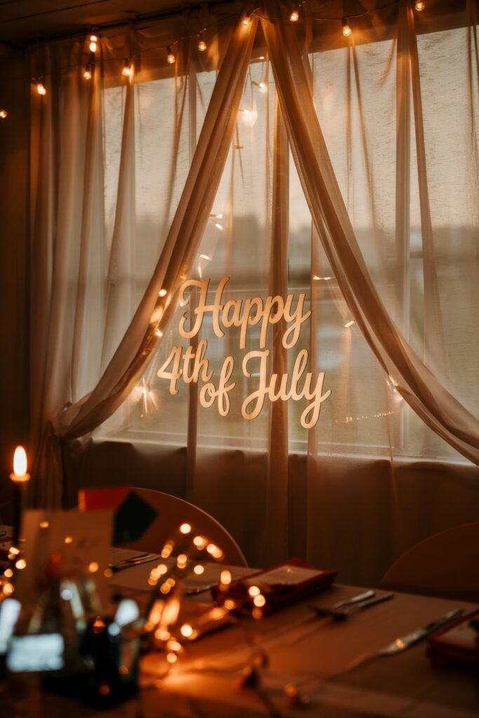 Patriotic 4th of July Table Décor & Centerpieces 34 Softly lit dining room window with string lights and sheer curtains featuring a “Happy 4th of July” sign for a cozy Independence Day atmosphere.