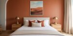 Minimalist bedroom with a terracotta accent wall, desert landscape artwork, and a white bed with rust and cream pillows, flanked by matching wooden nightstands and brass lamps, showcasing Terracotta Bedroom Ideas.