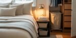 Cozy bedroom setup featuring a neatly made bed with soft neutral-toned pillows, a nightstand with a reading lamp and books, and an open closet area with a laundry hamper—ideal inspiration for bedroom essentials for first apartment.