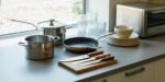 A countertop set near a window with kitchen essentials for a first apartment, including a saucepan, stockpot, skillet, cutting board with three knives, stacked plates and bowls, drinking glasses, and a toaster oven.
