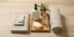 Neatly arranged bathroom essentials on a wooden surface, including a folded towel set, a rolled towel, a white soap dispenser, a reed diffuser, a potted plant, and a bar of soap in a dish, ideal for First Apartment Planning.