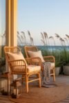 Two rattan chairs with white cushions and a striped blanket on a wooden deck overlooking the ocean, perfect for coastal summer decor.