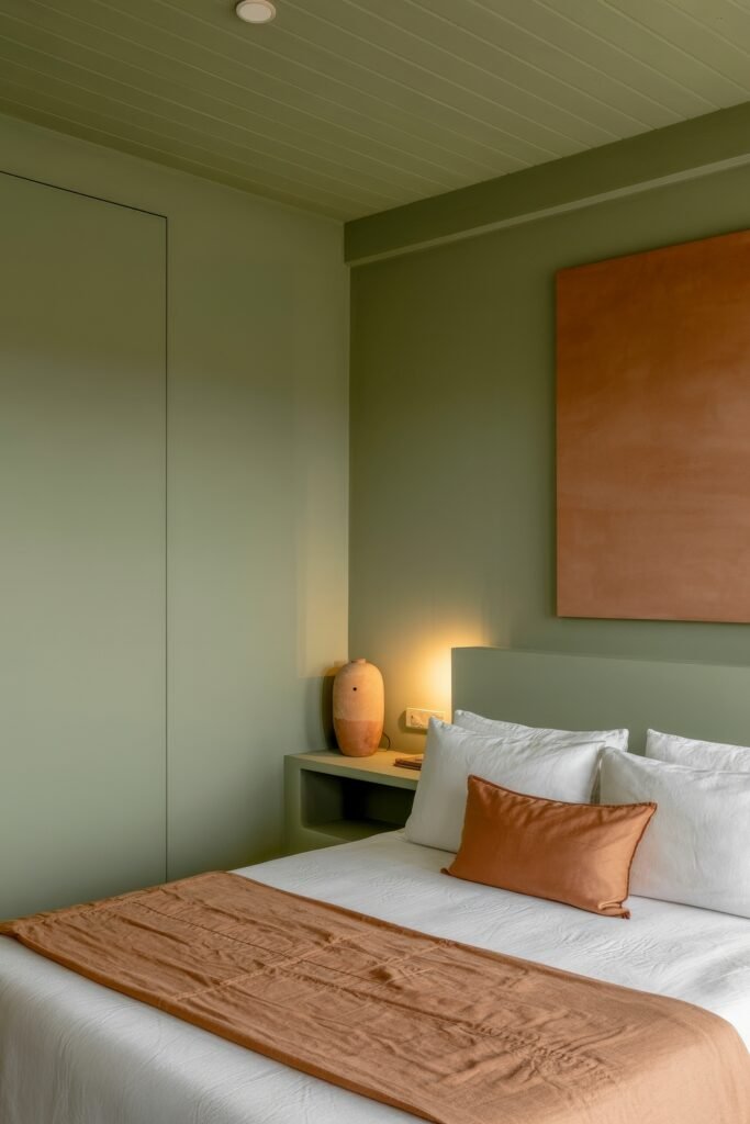 Earthy Bedroom Ideas: 12 Essential Ways to Elevate Your Space 4 Modern bedroom with green walls and ceiling, crisp white bedding, and burnt orange accents on a throw and pillow, complemented by a sculptural ceramic lamp.