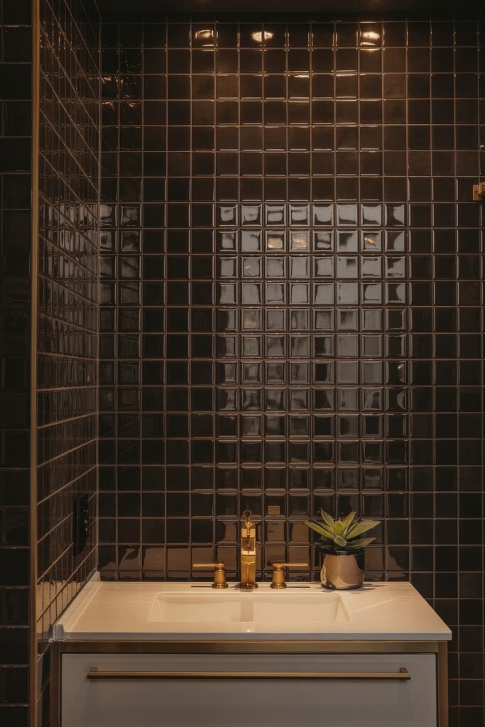 Black Bathroom Ideas: Genius Ways to Create Moody Glam 9 Glossy black tiled bathroom with golden faucet and hardware, a small green plant, and warm downlighting over a white vanity, blending luxury with a moody palette.