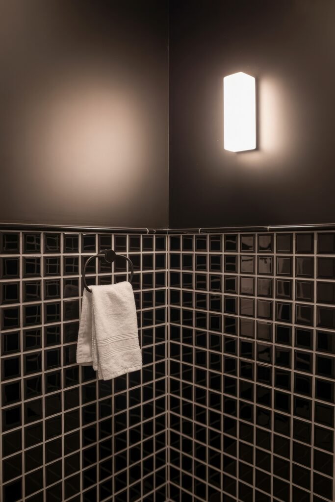 Black Bathroom Ideas: Genius Ways to Create Moody Glam 10 Corner of a bathroom with glossy black tiles, a white hand towel on a matte black ring holder, and a bright rectangular wall sconce, creating bold contrast in a dark space.
