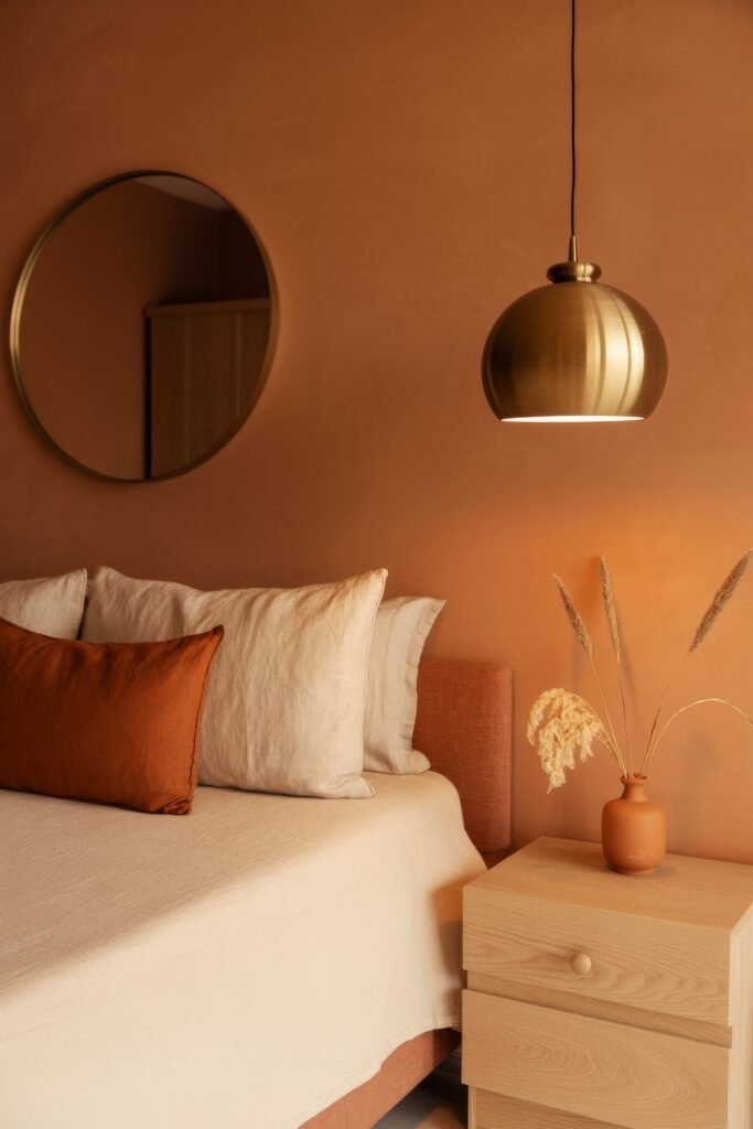 Terracotta Bedroom Ideas: Genius Ways to Glow 18 Minimalist bedroom with a rust-colored wall, linen bedding, oversized rust cushion, golden dome pendant light, and a light wood nightstand with a small terracotta vase of dried grasses.
