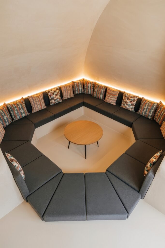 Sunken living room Magic: Cozy Tips for Instant Depth 3 Contemporary sunken seating area with dark gray cushioned benches arranged in a circular layout around a round wooden table, backed by ambient lighting and patterned pillows.