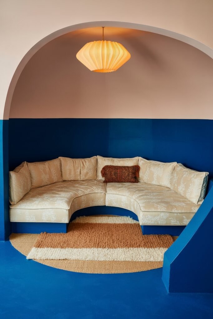Sunken living room Magic: Cozy Tips for Instant Depth 26 Bold sunken nook with a cream curved sectional placed under an arched alcove, featuring a blue-painted lower half, a striped rug, and a sculptural pendant lamp overhead.