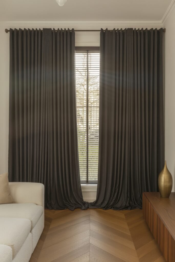 Luxury Curtains Living Room: Luxurious Curtains & Window Treatments 22 Minimalist interior featuring dark grey curtains around a central window with blinds, warm wood herringbone flooring, a plush cream sofa, and a sleek gold vase on a wooden media console.
