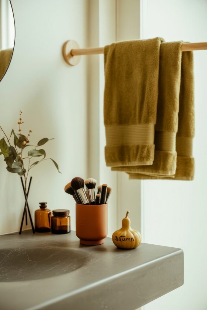 Fall Bathroom Decor: Cozy Upgrades for Instant Spa 30 Modern bathroom vanity with olive towels hanging from a wooden rod, amber glass jars, makeup brushes in a rust-colored holder, and a small gourd labeled "autumn," creating a playful and warm fall bathroom decor scene.