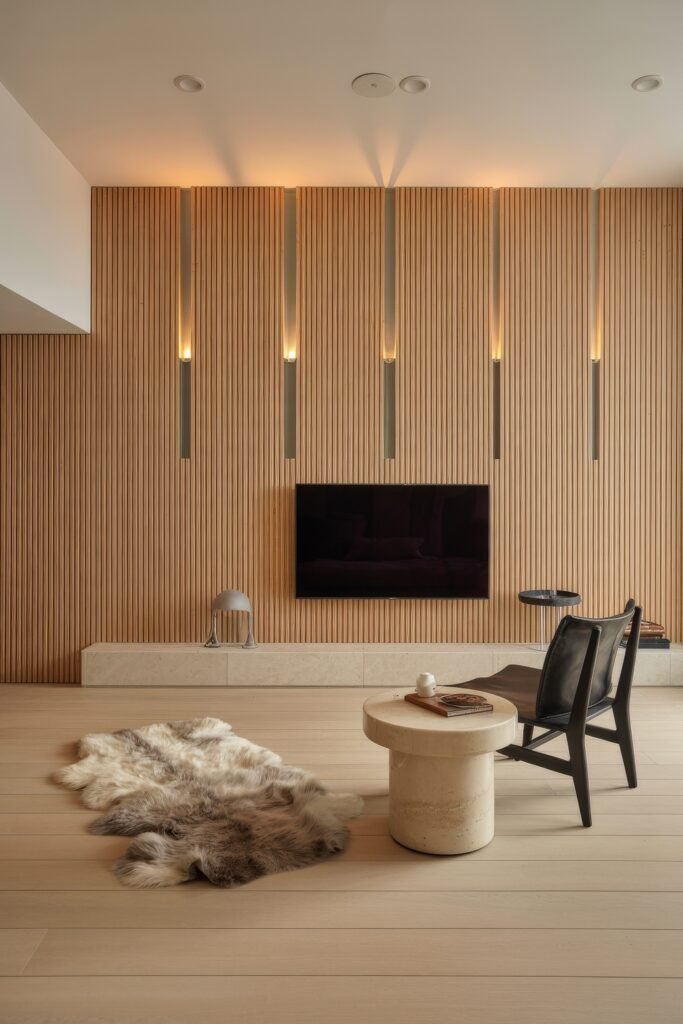 Living Room TV Wall Luxury: Sleek Ideas for a Stylish Setup 11 Minimalist luxury living room with a flat-screen TV mounted on a floor-to-ceiling vertical wood slat wall, accented by warm linear lighting and styled with a leather chair, round stone table, and soft fur rug.