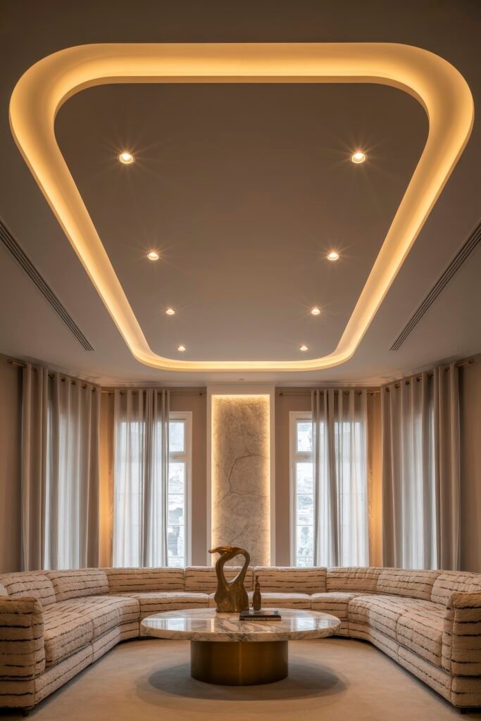 Luxury Ceiling Design in Living Rooms 9 Chic corner living room with panoramic windows, curved modular sofa, circular marble coffee table with bronze decor, and a softly glowing geometric recessed ceiling emphasizing the luxury ceiling design living room style.