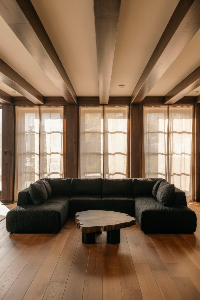 Luxury Ceiling Design in Living Rooms 12 Earthy-toned lounge featuring a black textured sectional sofa, organic-shaped wood coffee table, and exposed dark ceiling beams that contrast the soft filtered light from sheer curtains.