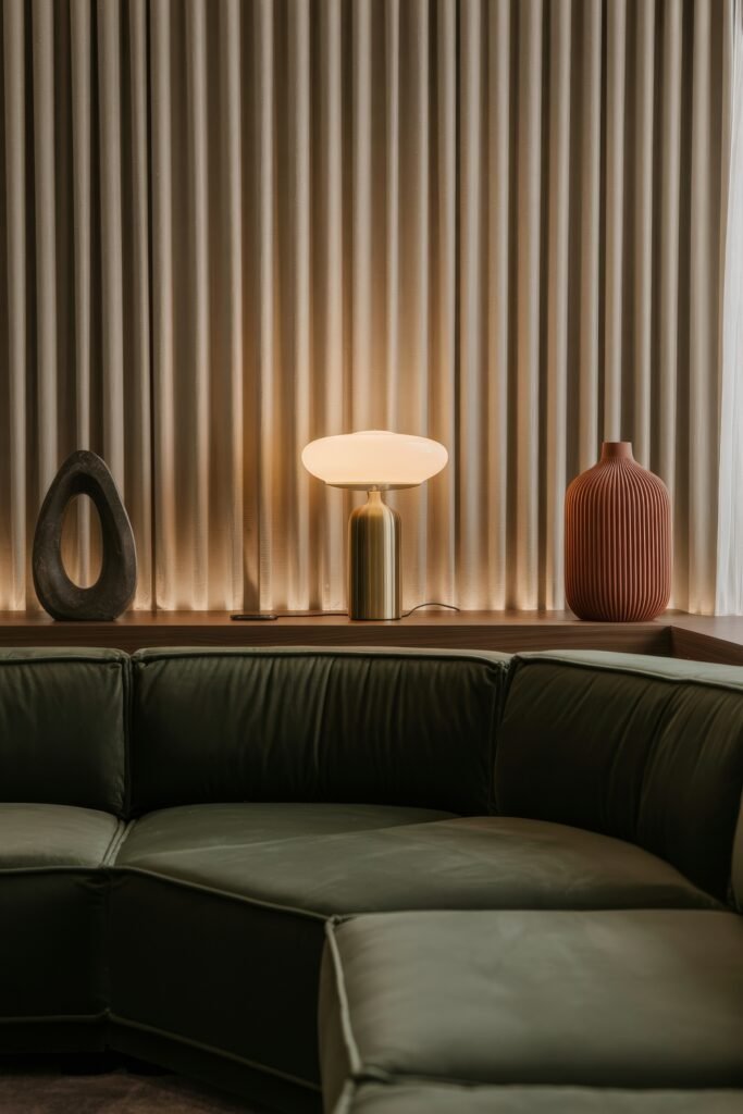 Modern Luxury Living Room: Designing a Sleek Luxe Lounge 18 Close-up of a dark green leather sectional backed by walnut shelving that holds a brass mushroom lamp and ribbed terracotta vase against pleated drapes