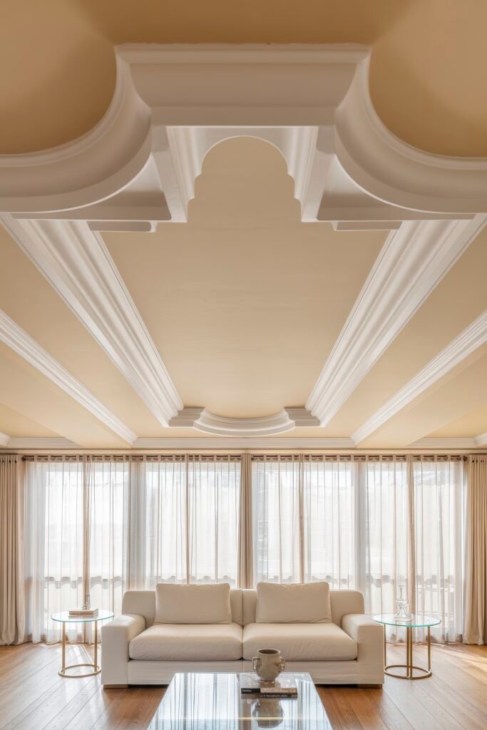 Luxury Ceiling Design in Living Rooms 20 Classic living room with ornate white ceiling moldings, full-length sheer curtains, and a neutral sofa ensemble, offering a graceful and airy ambiance with architectural charm.