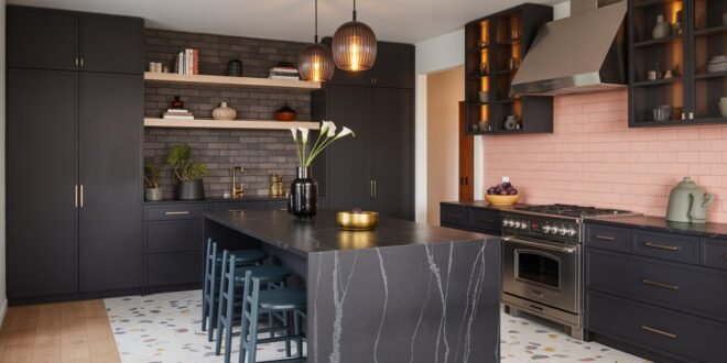 Spacious modern black kitchen with dark cabinetry, a black marble island, gold fixtures, and contrasting pink tile backsplash, lit by pendant lights over a terrazzo floor.