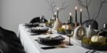 Elegant table set with black dinnerware, gold flatware, and black-and-white checkered napkins, featuring black candles, minimalist vases, and white pumpkins on moss centerpieces, creating a chic black and white thanksgiving tablescape.