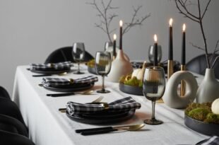 Elegant table set with black dinnerware, gold flatware, and black-and-white checkered napkins, featuring black candles, minimalist vases, and white pumpkins on moss centerpieces, creating a chic black and white thanksgiving tablescape.