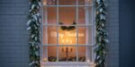 Festive window framed with snowy garlands and warm lights, displaying a “Merry Christmas” sign, a hanging wreath, and a glowing candleholder inside — a classic example of outdoor Christmas window decorations.