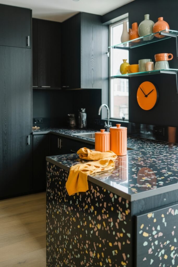 Chic Ways to Style a Modern Black Kitchen 28 Modern black kitchen with colorful terrazzo island and counters, orange and yellow accents, and glass shelving styled with pottery, illuminated by natural light.