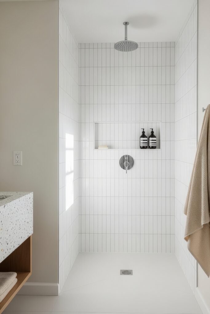 Creative Ways to Elevate Your Bathroom Wall Decor 4 Bright and minimalist walk-in shower with vertical white tile, a rain shower head, and recessed shelving holding black bottles, alongside a terrazzo vanity and beige towel on the wall.