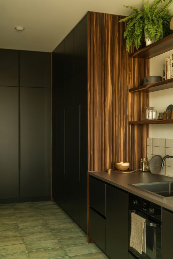 Chic Ways to Style a Modern Black Kitchen 7 Warm-toned modern black kitchen with vertical wood paneling, floating wood shelves, green tiled floor, and minimal countertop decor.