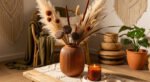 A terracotta vase filled with pampas grass and dried botanicals sits on a wooden coffee table beside a lit candle, creating a warm boho fall decor vibe.