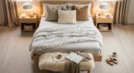 Soft neutral-toned cozy bedroom with a neatly made bed layered in cream bedding, textured knit pillows, and a plush bench topped with an open book, tea cup, and throw blanket.