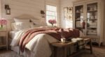 Sweet Ideas for a Romantic Cozy Farmhouse Bedroom Sunlit romantic cozy farmhouse bedroom with a cream upholstered bed layered in soft floral bedding, a chunky pink knit throw, fresh pink flowers, and a wooden bench set with a cup of coffee.