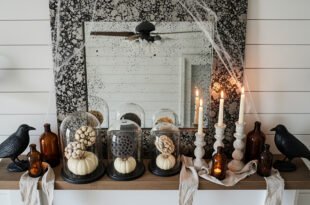 Rustic mantel decorated with faux cobwebs, black crow figurines, amber glass bottles, pillar candles, and cloche-covered white pumpkins for farmhouse Halloween home décor.