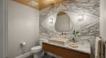 Modern half bathroom with a marble accent wall, floating wood vanity, vessel sink, and abstract mirror framed by two wall sconces.