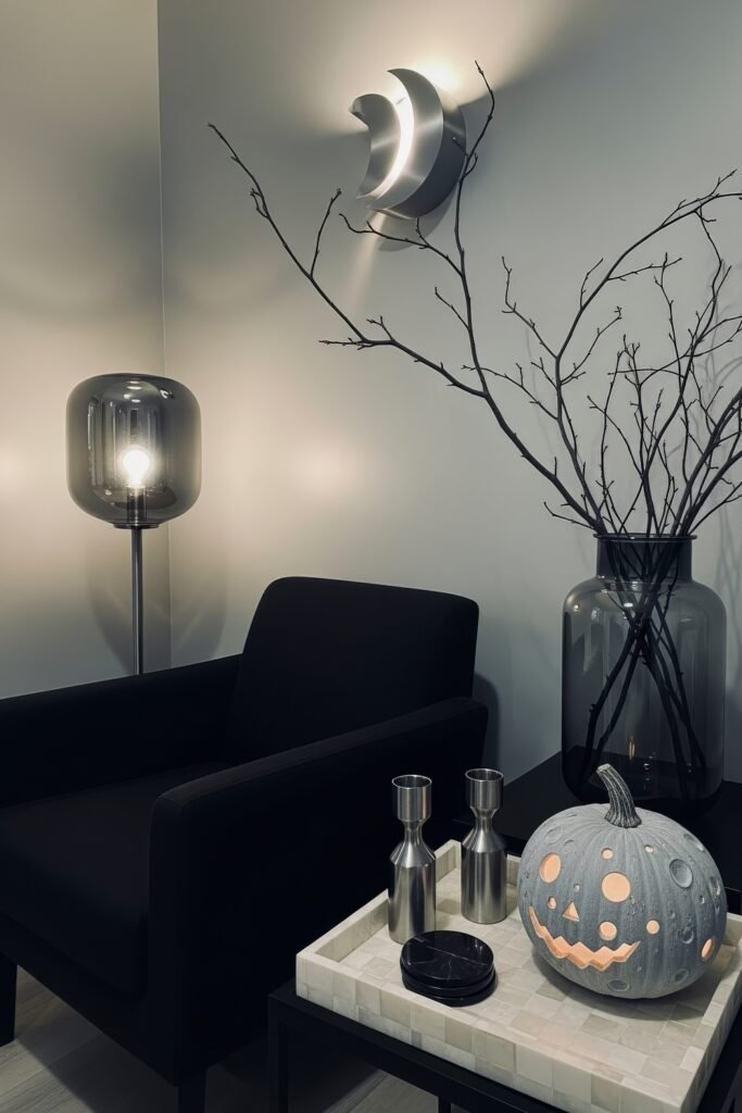 Spooky-Chic Magic: halloween home décor Ideas You’ll Love 10 Minimalist corner with a gray moon-textured pumpkin lantern, black armchair, bare branches, and modern lighting shaped like a crescent moon.