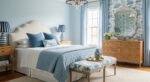 Fresh blue bedroom ideas for a calm, chic retreat Elegant bedroom with light blue walls, white upholstered headboard, patterned bench, and layered blue bedding accented by striped lamps and wicker furniture.