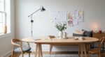 Bright dining room office combo with a wooden oval table styled with books, a laptop, and a vase of greenery, set against a wall-mounted inspiration board and neutral seating.