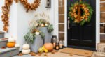 Front porch decorated with a black door wreath of greenery, small pumpkins, and oak leaves, surrounded by stacked pumpkins, lanterns with candles, and leafy garlands for a warm Thanksgiving Décor look.