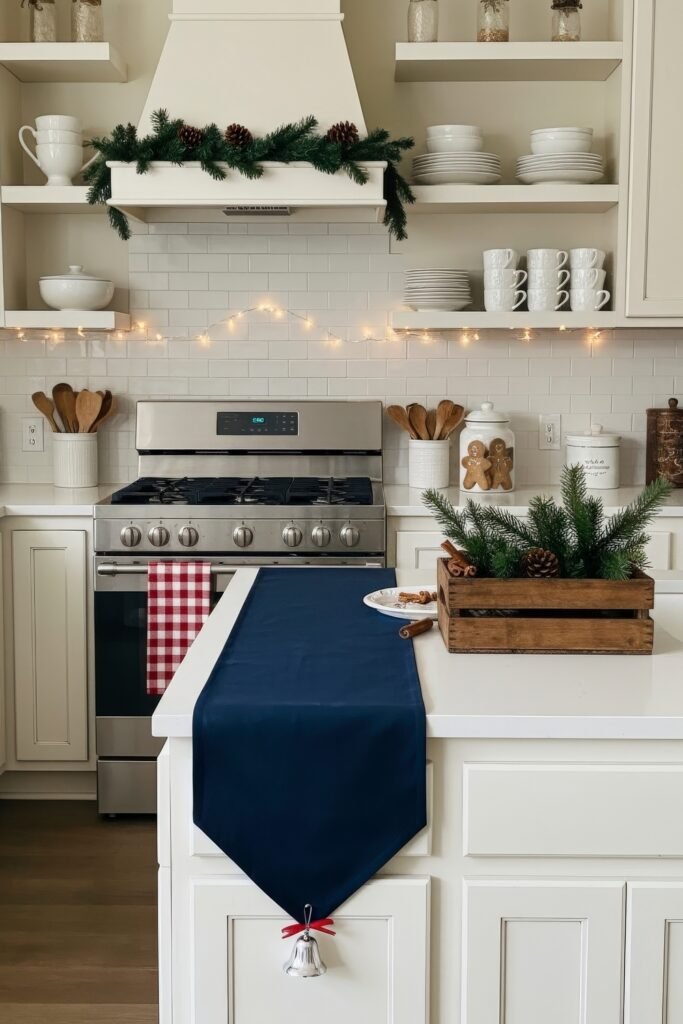 Cozy Ways to Style a Christmas Kitchen That Feels Magical 16 Traditional kitchen decorated with pine garland, twinkle lights, and pinecones, featuring open shelves of white dishes and a navy runner with a bell on the island.