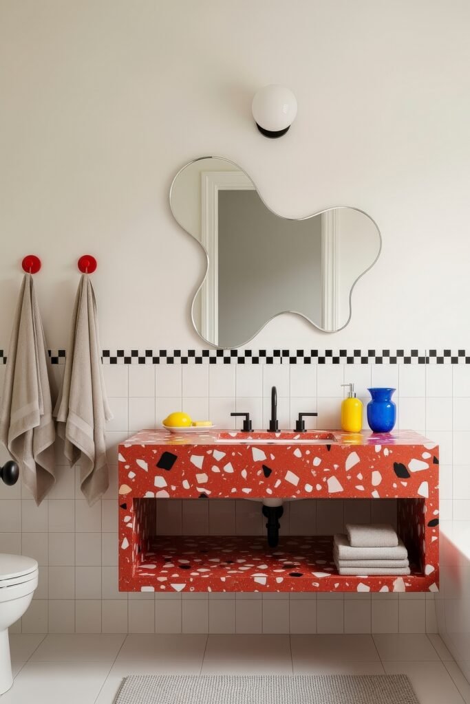 13 Bold Ideas for Red Bathroom Decor That Wow 11 Playful bathroom with a bold red terrazzo vanity, wavy mirror, and colorful accents in yellow and blue against white tile walls.