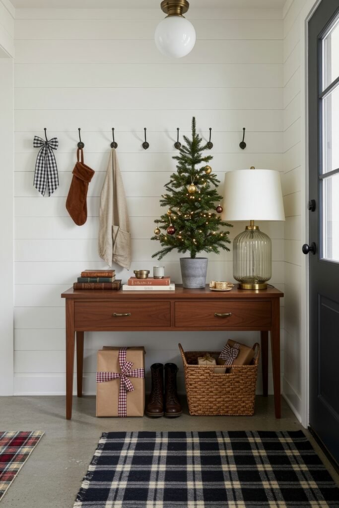 Cozy Christmas Décor for a christmas entryway 12 A simple christmas entryway with a wood console holding a potted mini tree, books, and a glass lamp, paired with wrapped gifts, boots, and a woven basket below.