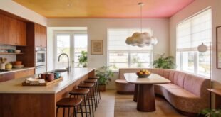 Warm and inviting kitchen dining room combo featuring wood cabinetry, a long island with bar stools, and a curved banquette with a wood table under a cloud-shaped pendant light, surrounded by natural light and houseplants.