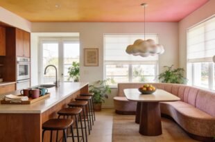 Warm and inviting kitchen dining room combo featuring wood cabinetry, a long island with bar stools, and a curved banquette with a wood table under a cloud-shaped pendant light, surrounded by natural light and houseplants.