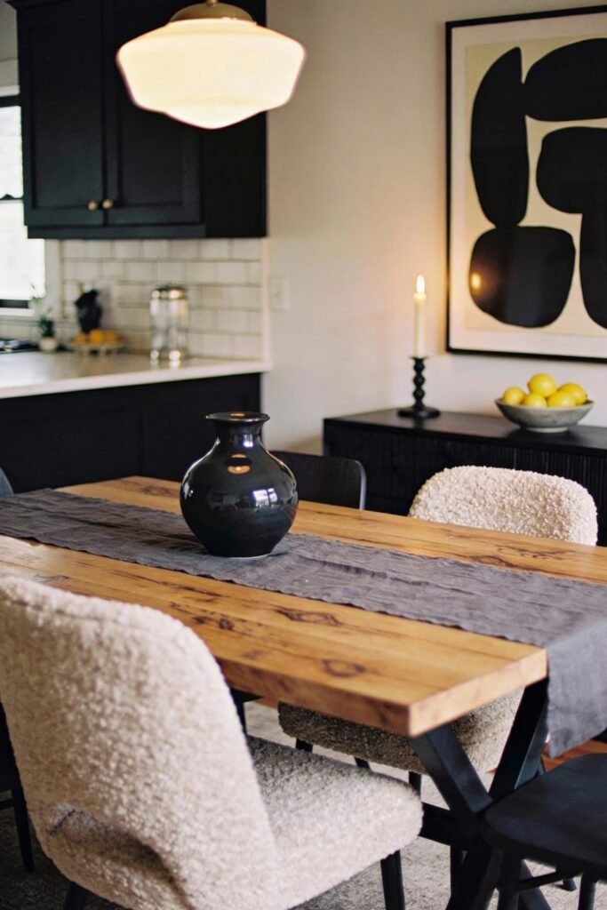 Kitchen Dining Room Combo Ideas That Feel Effortless 13 Moody kitchen dining room combo featuring a rustic wood table with shearling chairs, matte black cabinets, and bold abstract wall art, accented by candlelight and minimal decor.