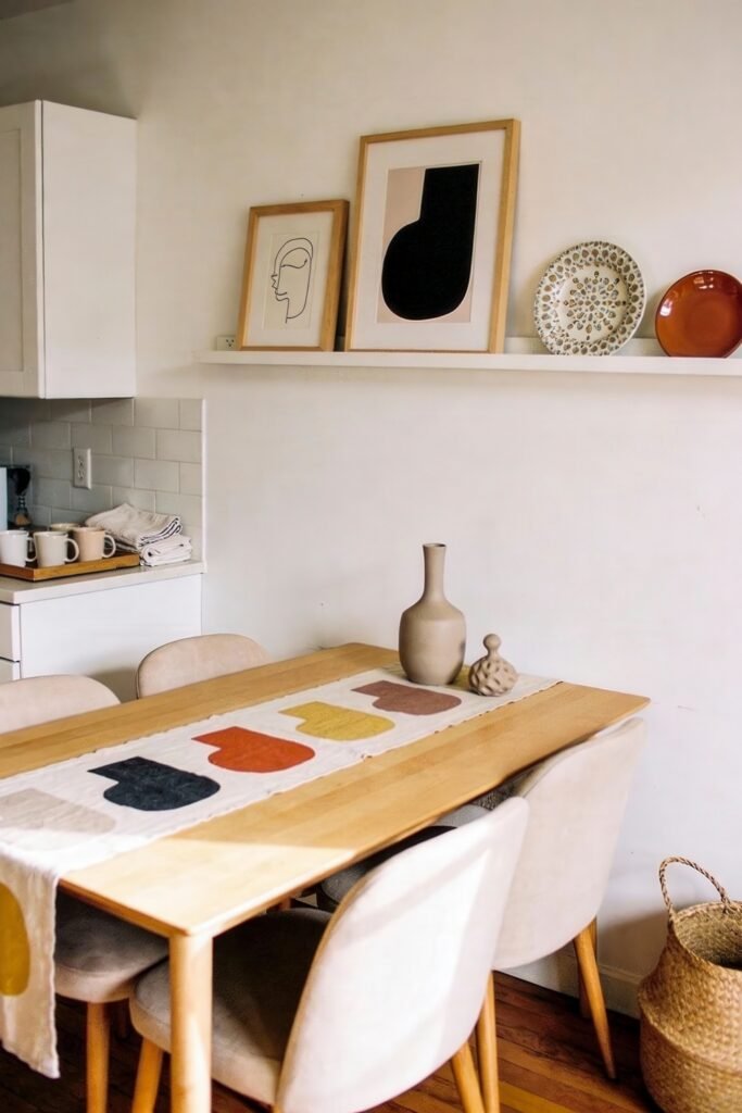 Kitchen Dining Room Combo Ideas That Feel Effortless 18 Minimalist kitchen dining combo with pale wood furniture, soft upholstered chairs, abstract wall art on a shelf, and a simple tablescape, all in a calm, neutral-toned setting.