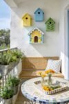 Cute Birdhouse Painting Ideas for Charming Outdoor Style Sunny balcony with colorful birdhouses on a white wall beside a wicker bench mosaic table yellow flowers and potted herbs