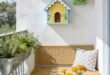 Cute Birdhouse Painting Ideas for Charming Outdoor Style 155 Sunny balcony with colorful birdhouses on a white wall beside a wicker bench mosaic table yellow flowers and potted herbs