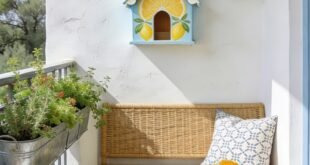 Sunny balcony with colorful birdhouses on a white wall beside a wicker bench mosaic table yellow flowers and potted herbs