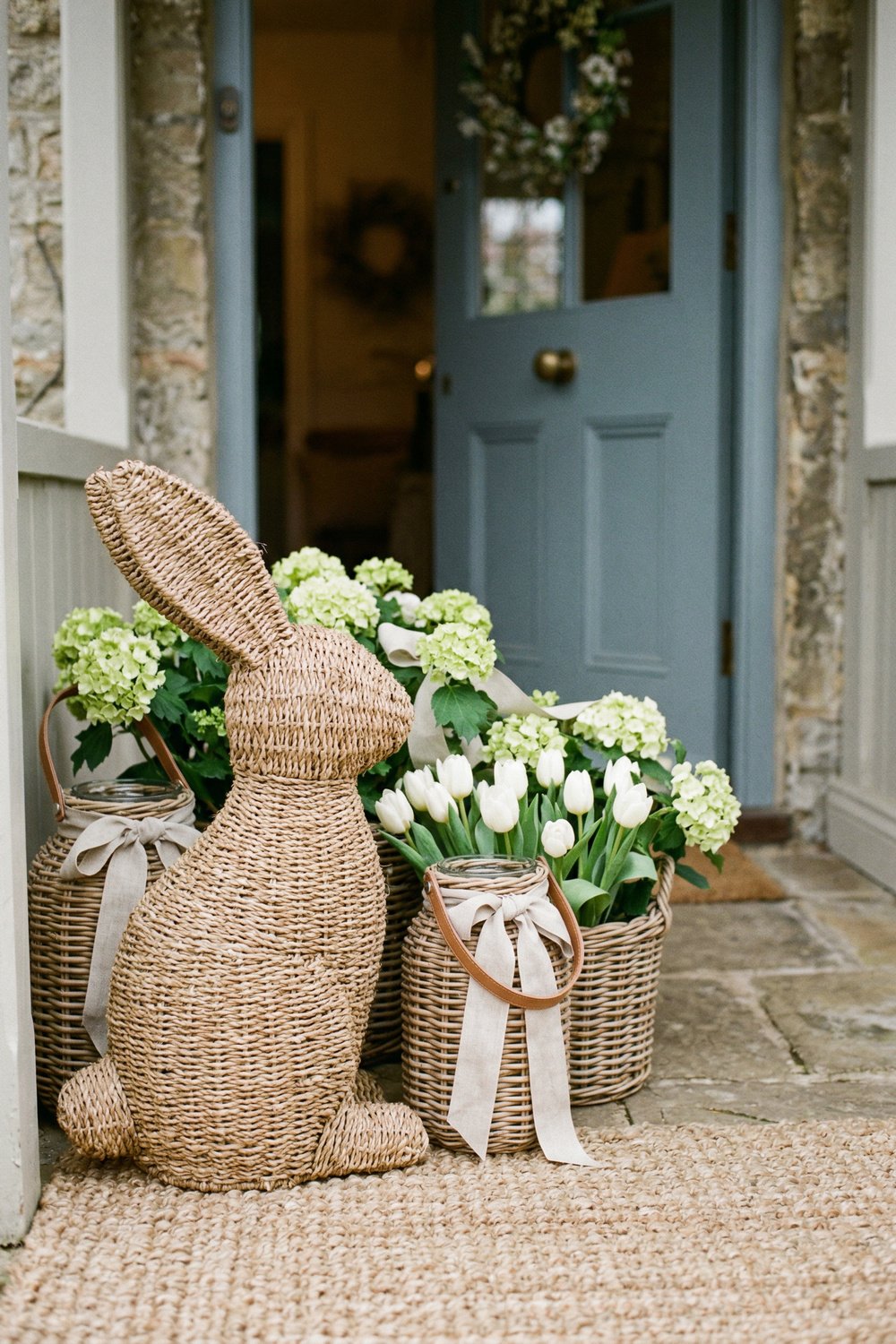 Woven bunny porch decor beside baskets of white tulips and hydrangeas at a blue front door