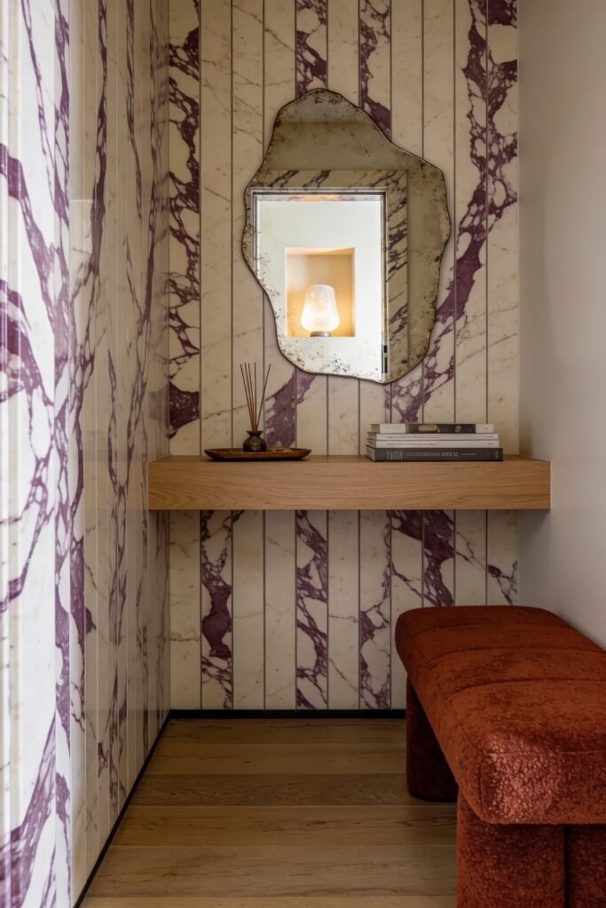 Small marble lined nook with a floating wood shelf antique mirror stacked books and a rust colored bench by a window