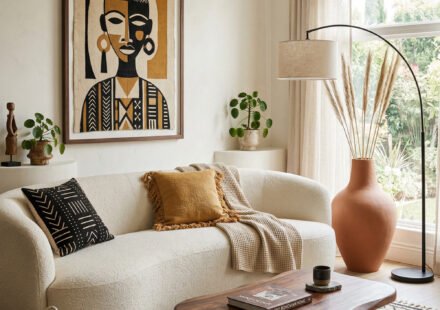 Curved cream sofa with ochre pillows, abstract portrait wall art, terracotta floor vase, arc lamp, and geometric rug in a warm Afro Boho Decor living room.